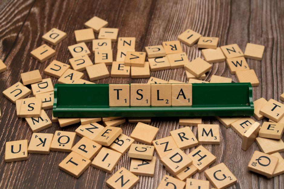 Wooden Scrabble tiles scattered on a wooden table with a green tile rack displaying the letters TSLA in order.